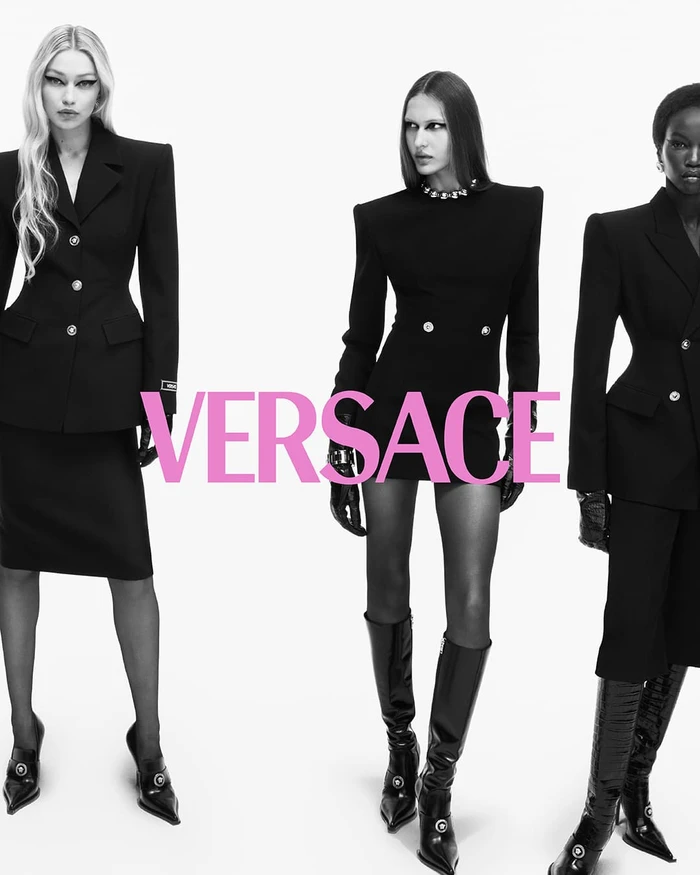 Three models wearing structured black tailoring and knee-high boots against a plain white background with bold pink Versace typography