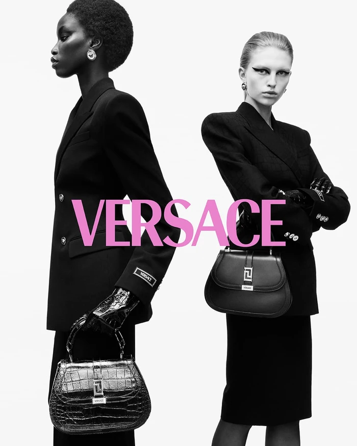 Two models wearing black blazers and holding black handbags against a plain white background with bold pink Versace text overlay.