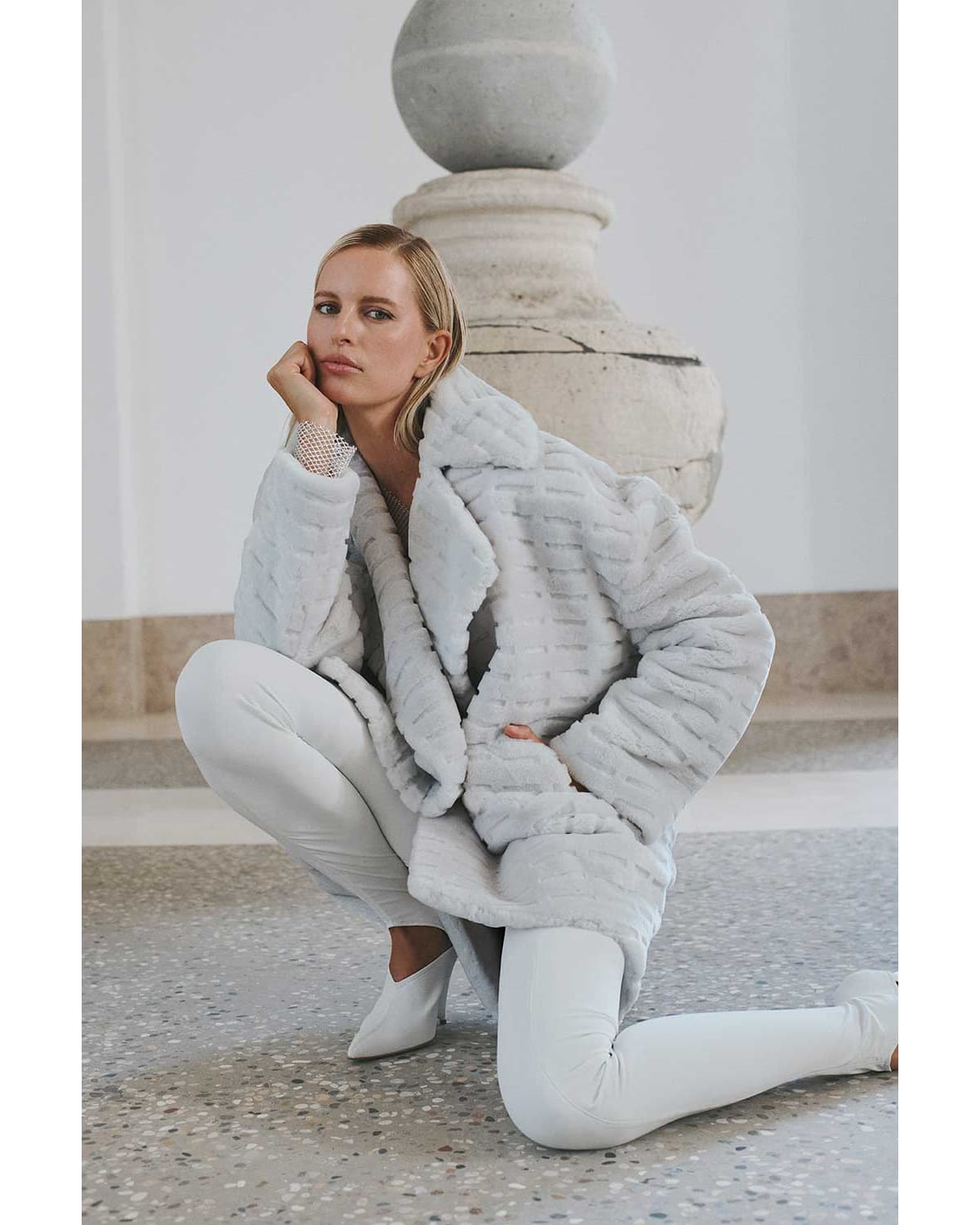 Fashion model in a crouched pose, wearing a light grey faux fur coat, paired with matching pants and boots, in a light-filled interior.