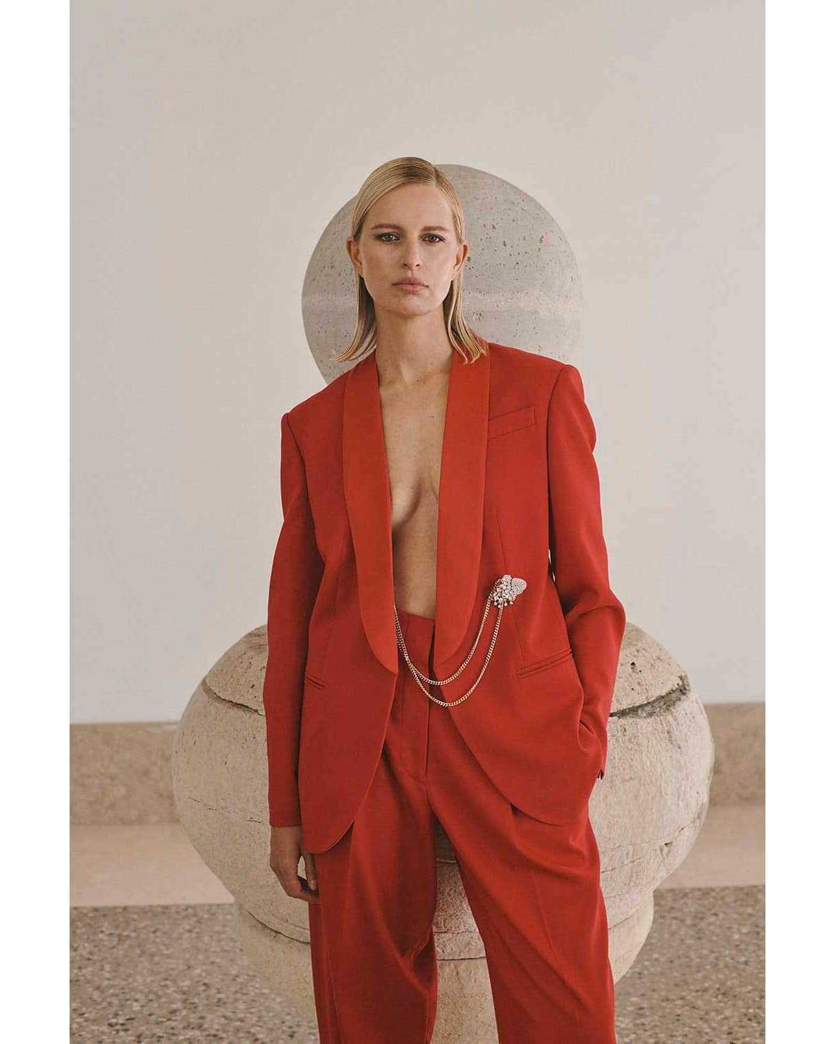 Woman in a red suit stands in front of a decorative stone pillar, posing in a sleeveless red suit with minimal jewelry, editorial fashion.