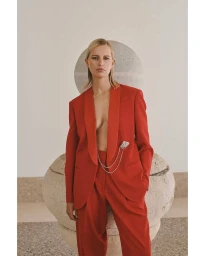 Karolina Kurkova posing in a vibrant red pantsuit with a silver chain brooch against a minimalist stone backdrop.