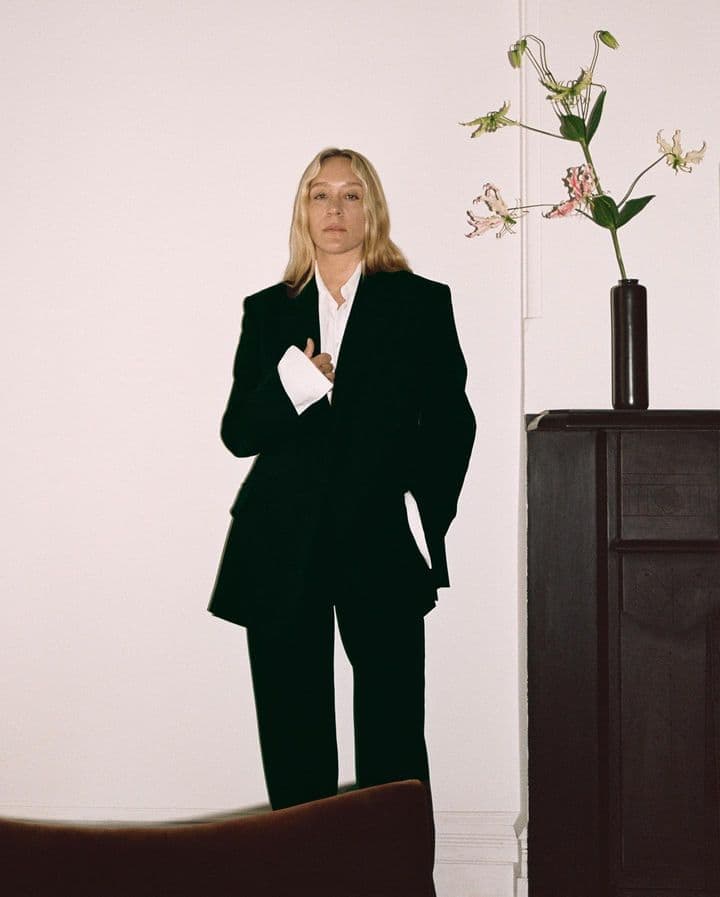 An adult female in a black suit and white shirt poses indoors, next to flowers and a dark cabinet, creating a stylish, modern portrait.