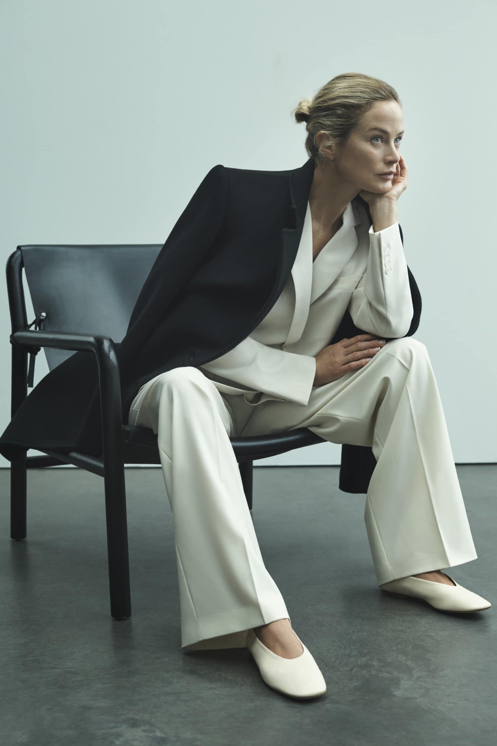 A woman sits in a black chair wearing a cream-colored pantsuit and black jacket in a studio, striking an elegant pose with a serene expression.