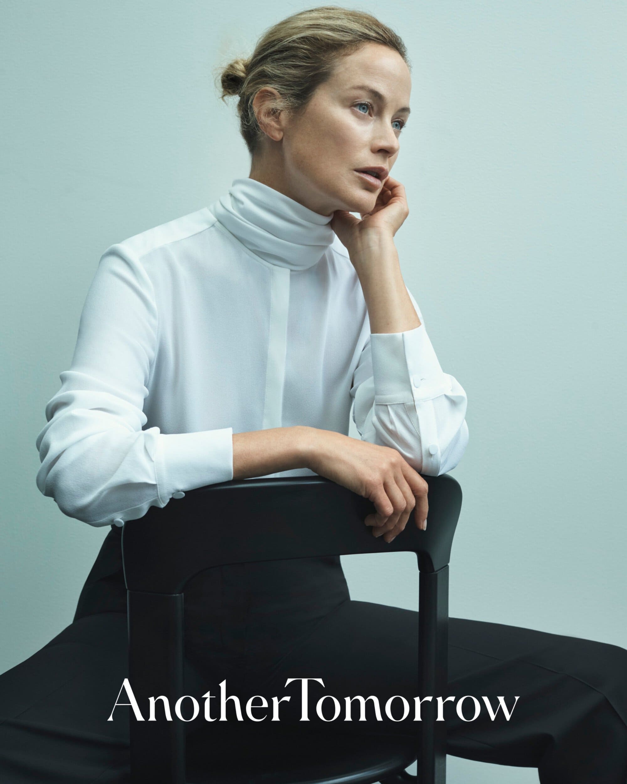 Blonde woman poses sitting on black chair in white turtleneck and black trousers in a studio setting for fashion brand ad.