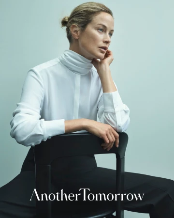 Blonde woman poses sitting on black chair in white turtleneck and black trousers in a studio setting for fashion brand ad.