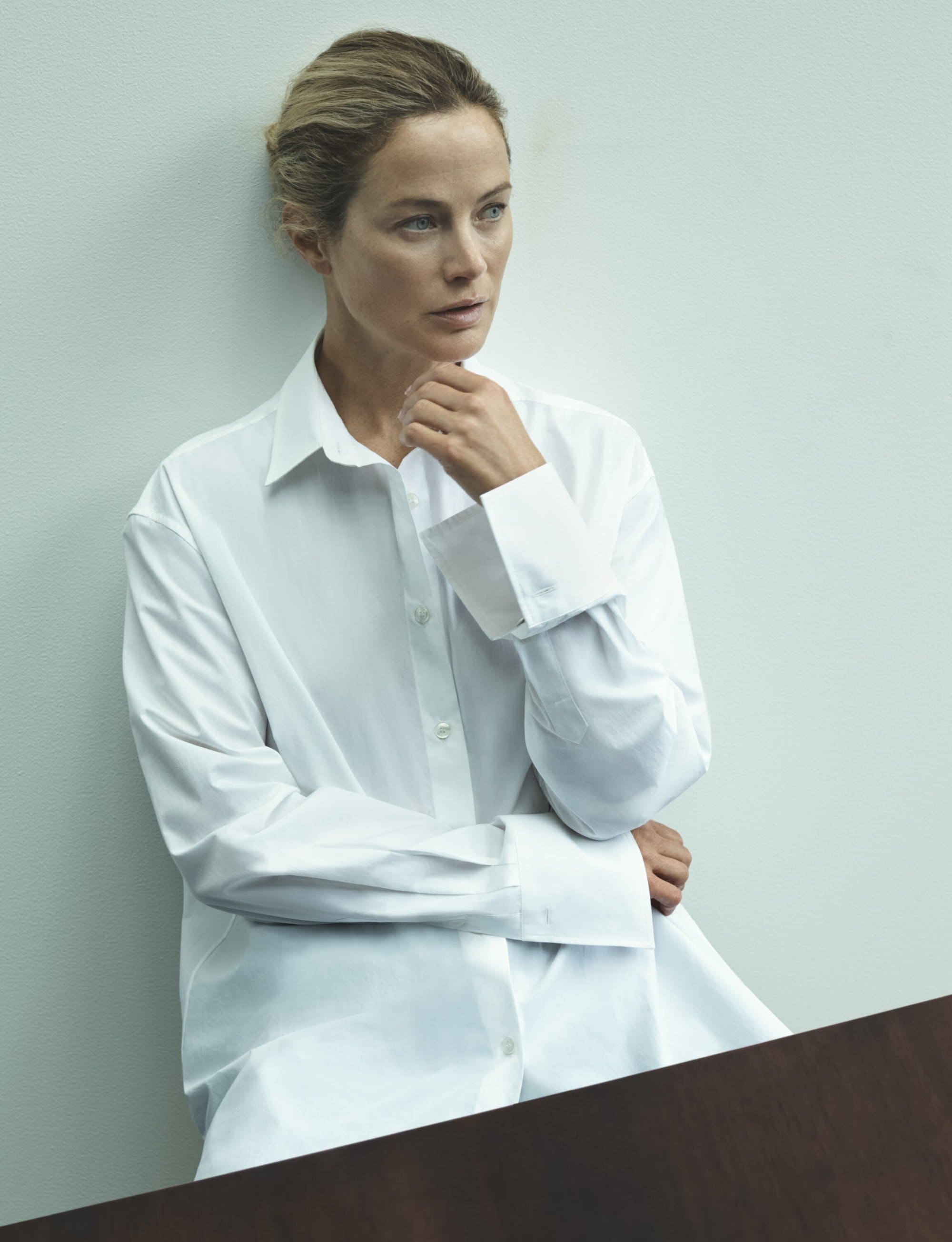 Elegant portrait of a mature woman in a white shirt leaning against a wall, showcasing minimalist fashion and serene expression.