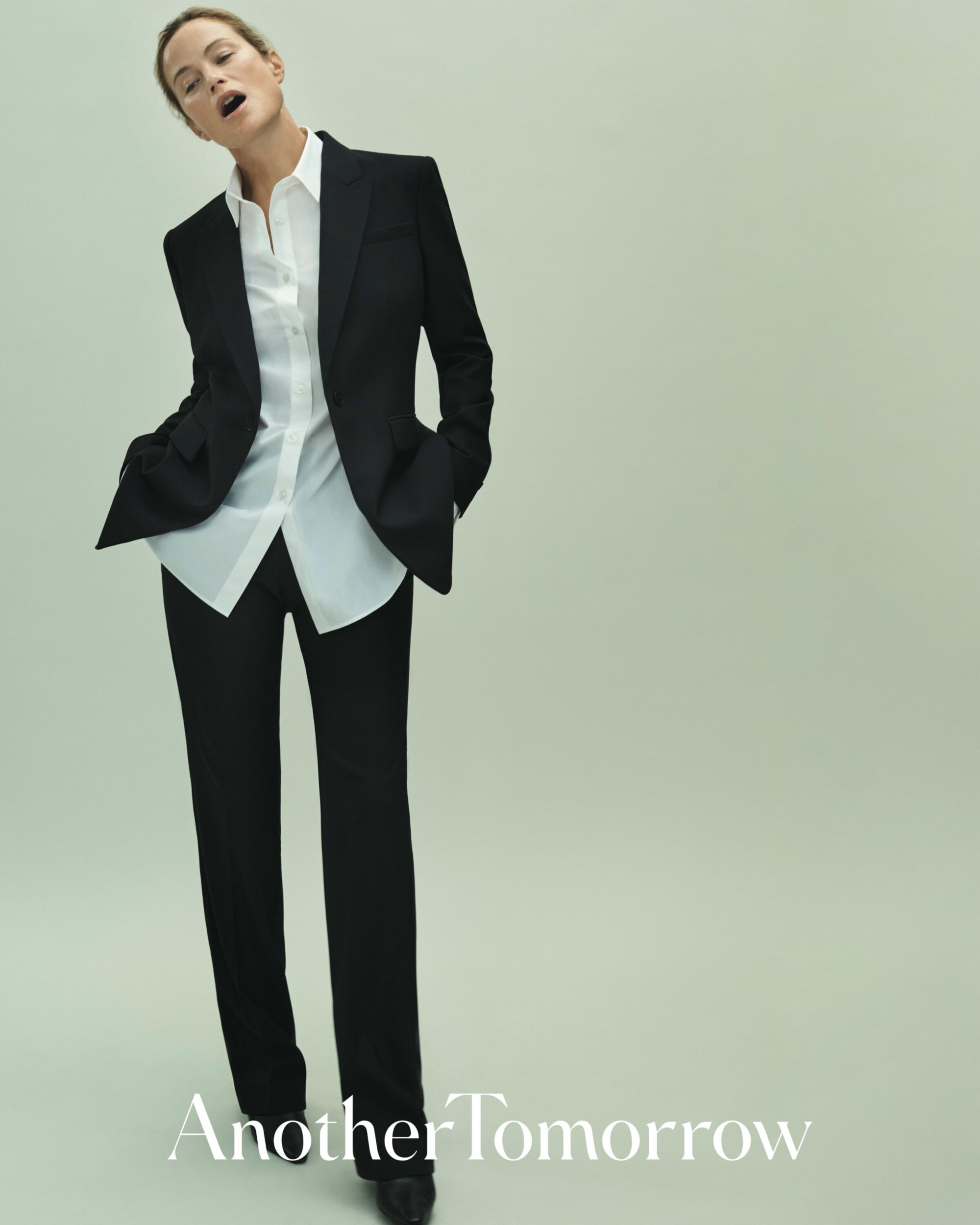 A full-length portrait of an adult woman in a black pantsuit and open white shirt, posing in a light-green studio setting.
