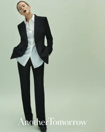 A full-length portrait of an adult woman in a black pantsuit and open white shirt, posing in a light-green studio setting.