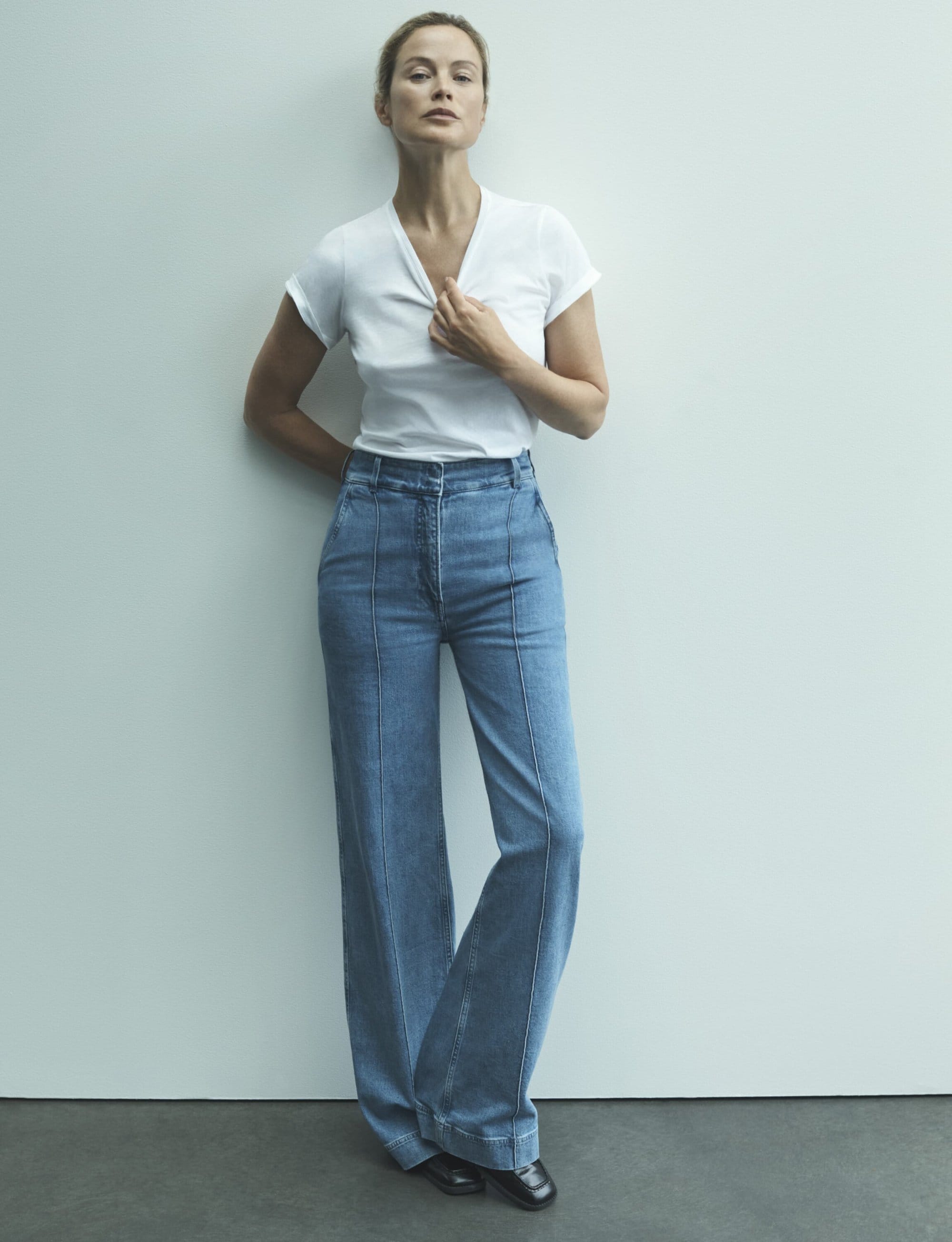 Full-length studio shot of a fair-skinned adult woman wearing a white t-shirt and light blue denim bell-bottom jeans, leaning against a white wall.