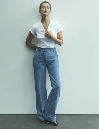 Full-length studio shot of a fair-skinned adult woman wearing a white t-shirt and light blue denim bell-bottom jeans, leaning against a white wall.