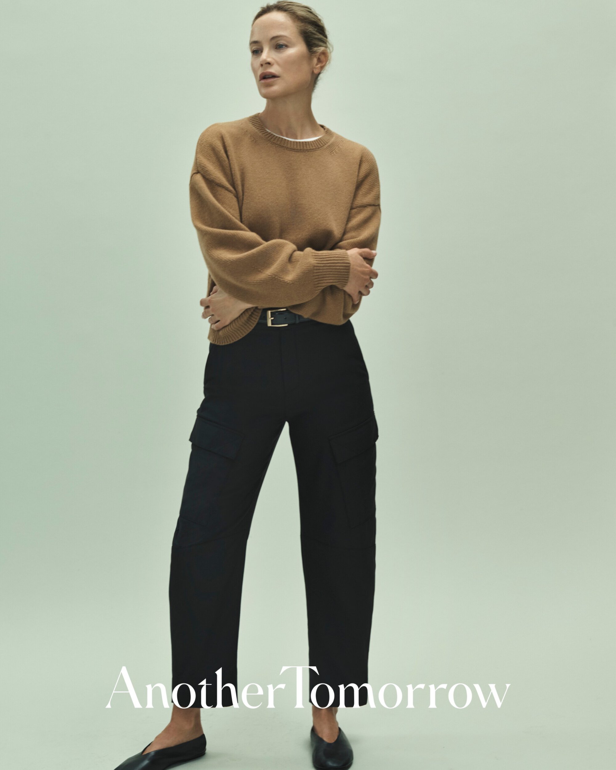 Fashion shot of a slender woman in a brown sweater and black pants, posed in front of a light green background, showcasing modern style.