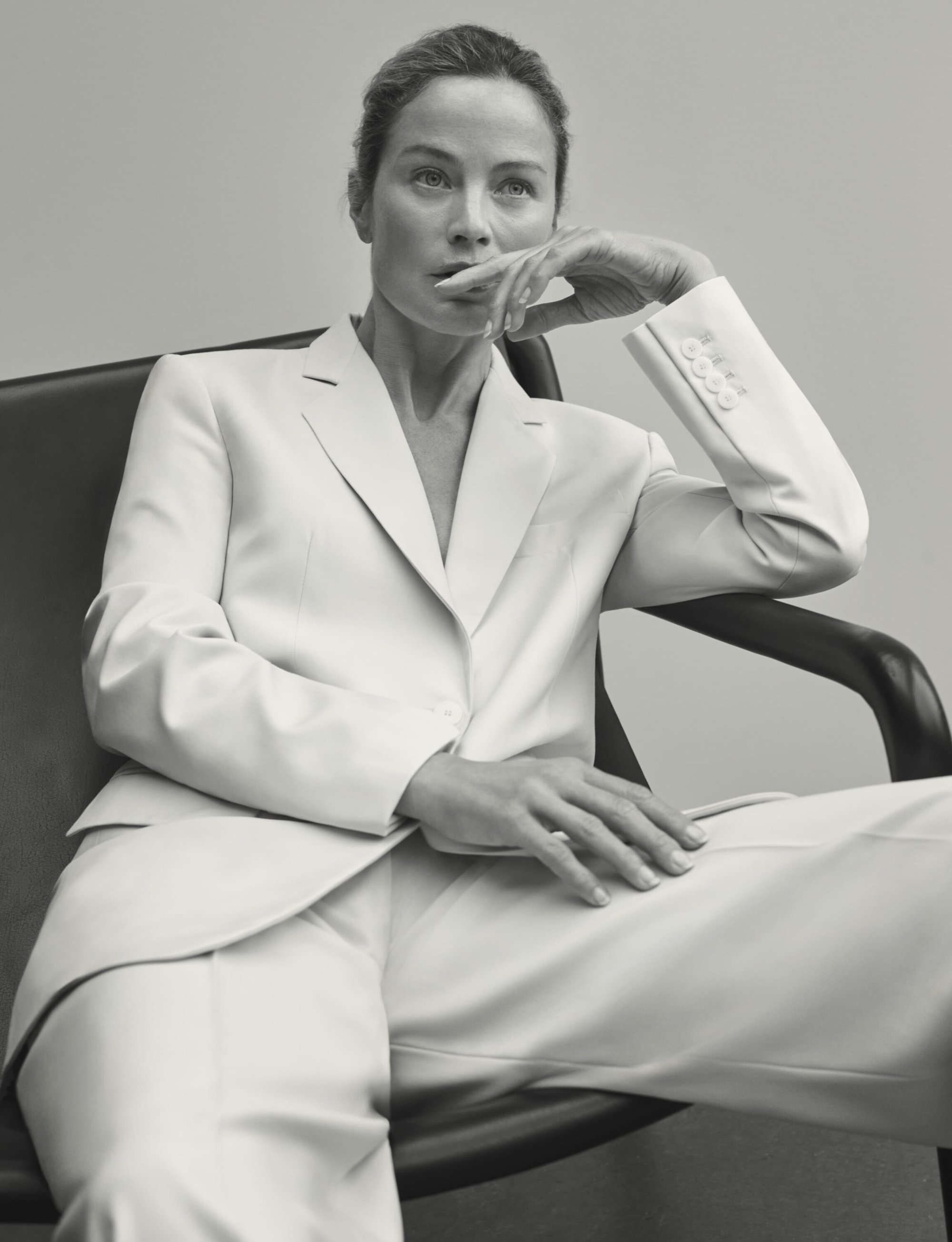 Portrait of a mature woman in a white suit sitting in a chair against a neutral background, looking thoughtfully to the side.