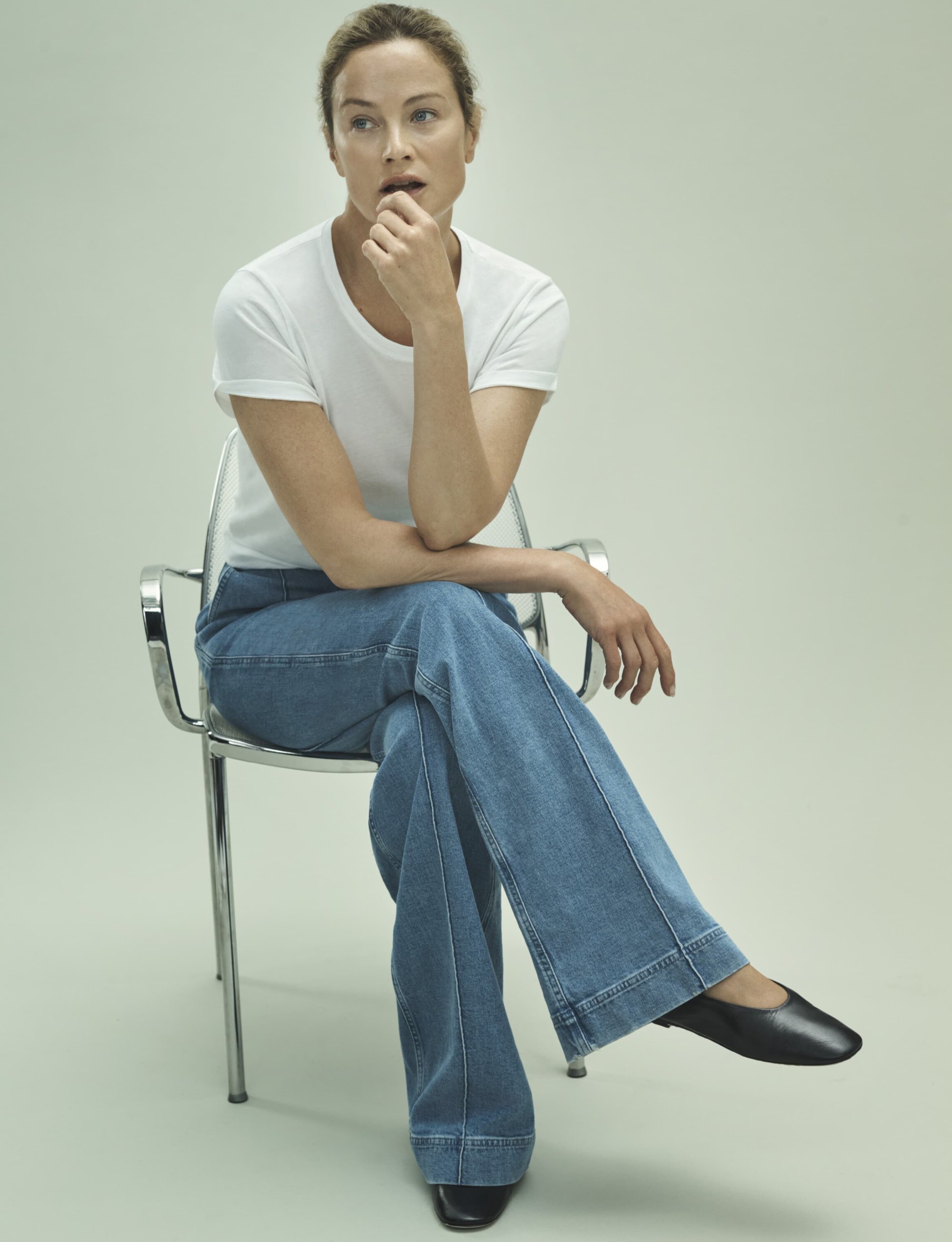 Adult woman sits on a clear plastic chair, wearing a white t-shirt and light blue jeans with black shoes, in a bright, neutral setting.