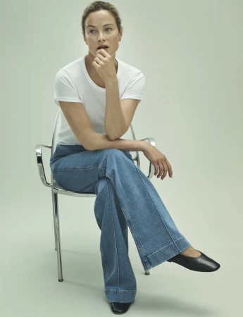 Adult woman sits on a clear plastic chair, wearing a white t-shirt and light blue jeans with black shoes, in a bright, neutral setting.