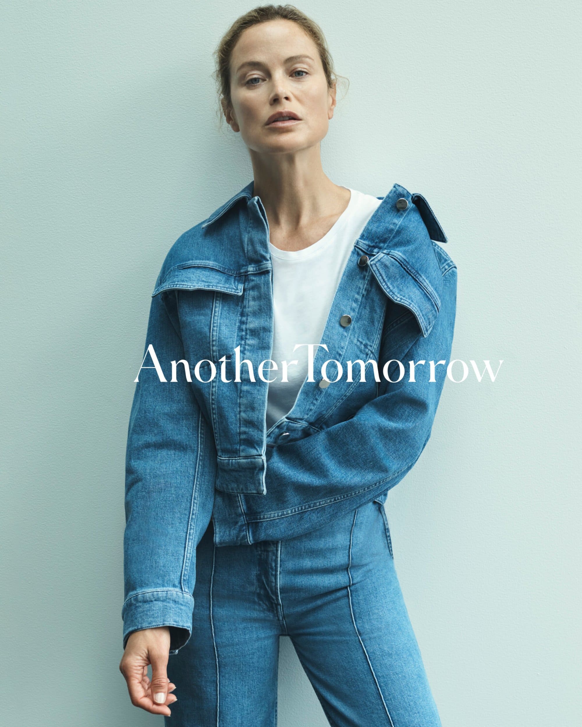 Fashion shot of a woman in a denim jacket and jeans against a light blue background. She has a neutral expression and casual pose.
