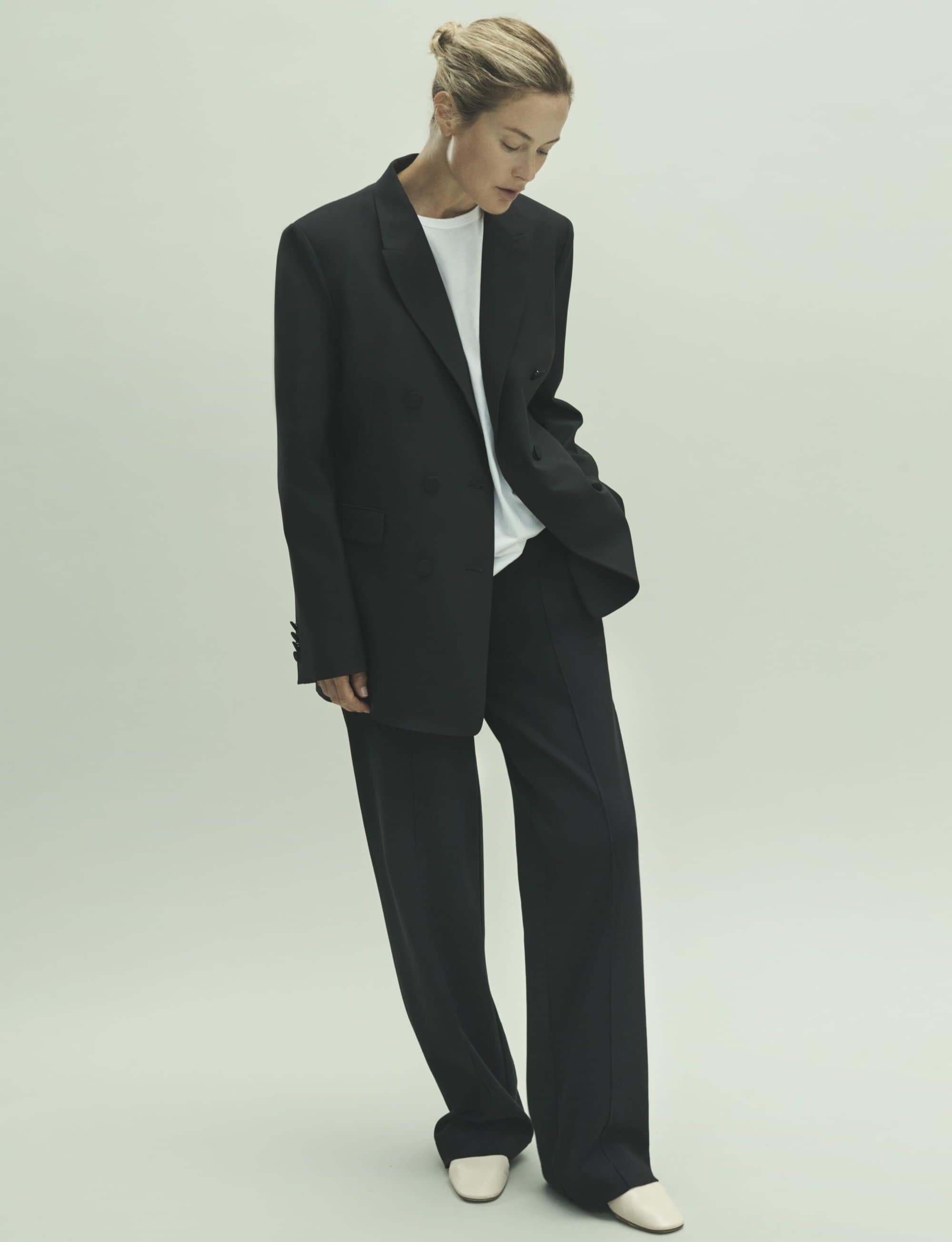 A woman posing in a studio wearing a black blazer, white t-shirt, and black pants in a fashion editorial shot.