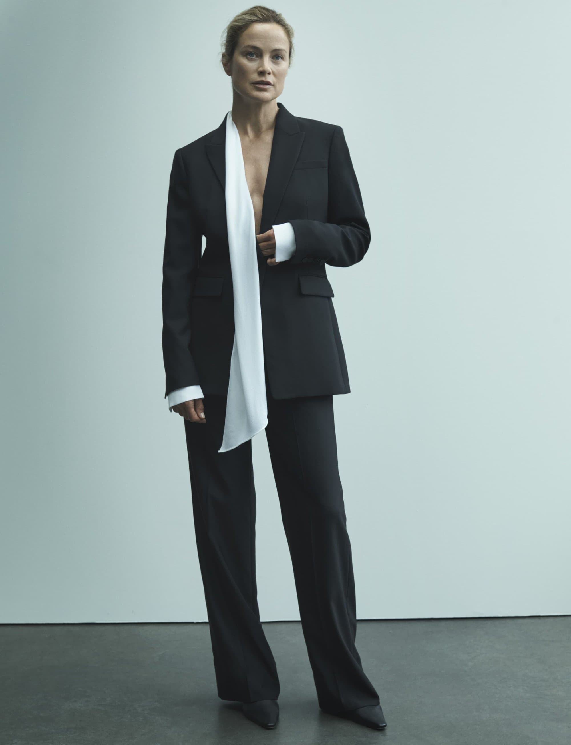 Model Carolyn Murphy standing in a tailored black trouser suit with a draped white scarf against a plain studio wall.