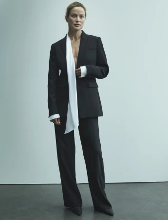 Model Carolyn Murphy standing in a tailored black trouser suit with a draped white scarf against a plain studio wall.