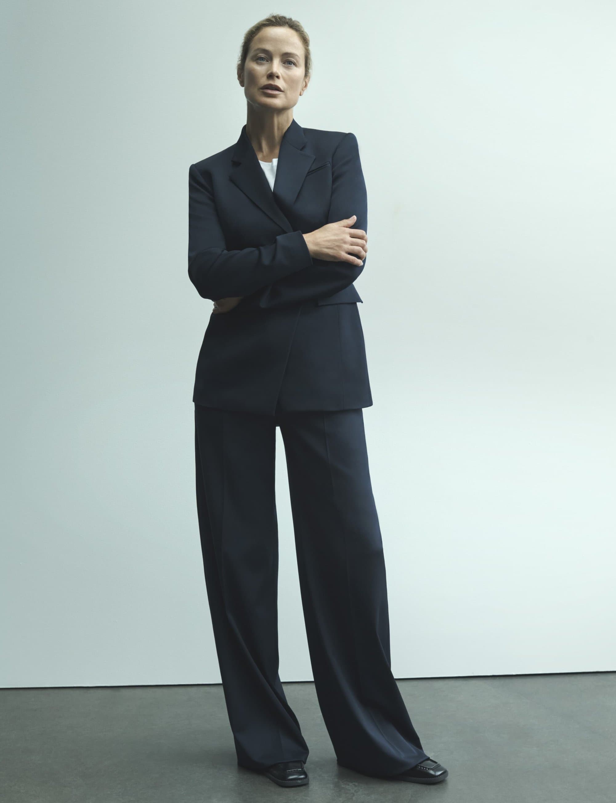 Elegant woman in a dark navy pantsuit stands confidently indoors against a light wall, embodying modern professional fashion.