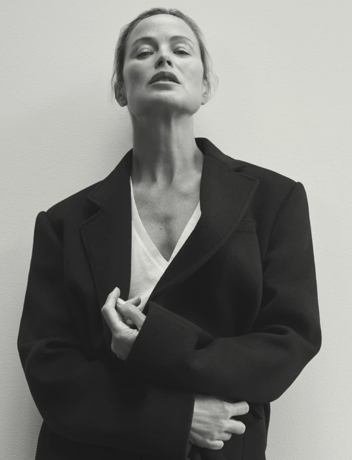 Carolyn Murphy posing in an oversized black tailored coat over a white v-neck top against a plain studio background.