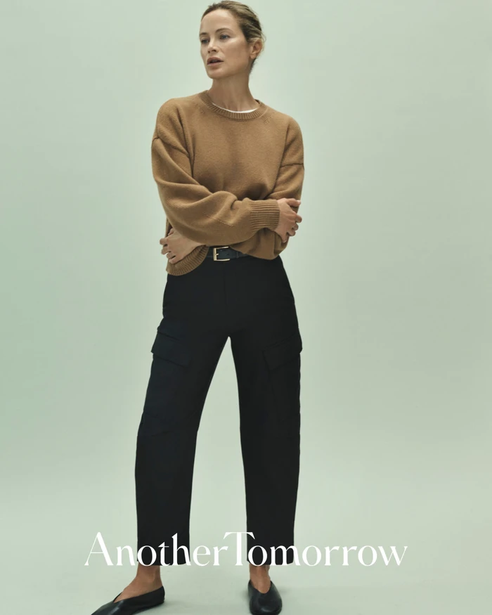 Model Carolyn Murphy wearing a camel knit sweater and black trousers against a pale green studio background.