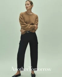 Model Carolyn Murphy wearing a camel knit sweater and black trousers against a pale green studio background.