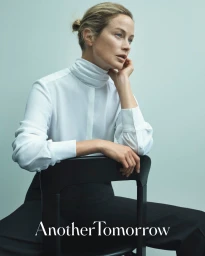 Model Carolyn Murphy wearing a white turtleneck blouse, seated on a black chair against a pale background