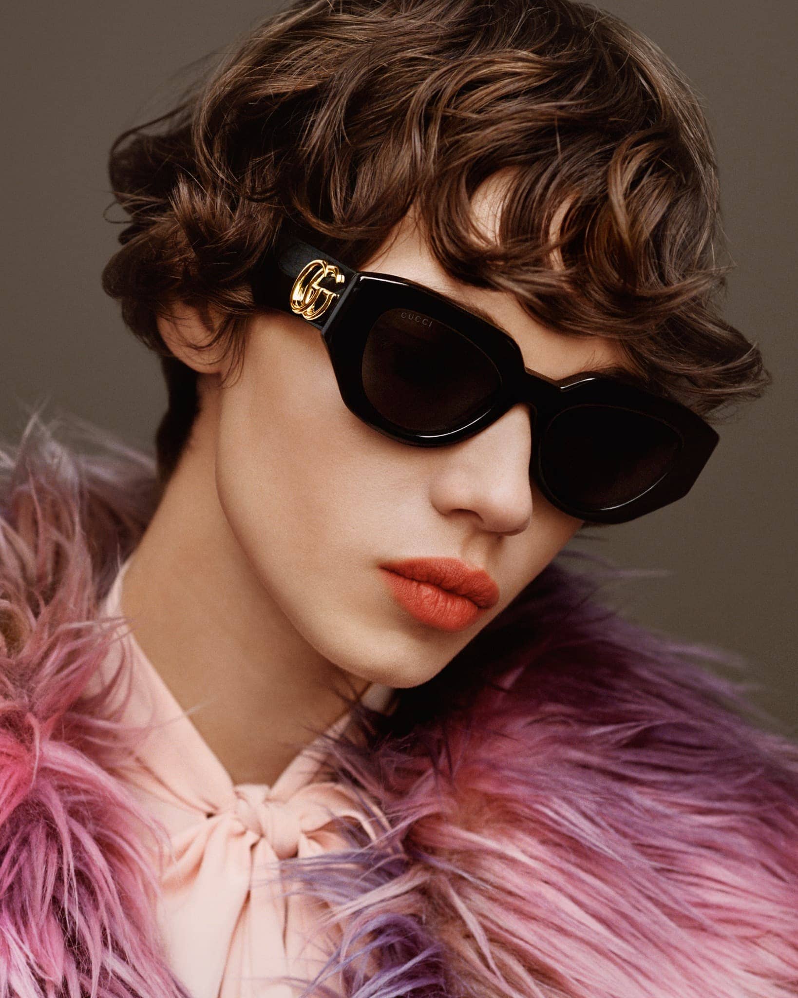 A woman with dark curly hair poses wearing black sunglasses with the Gucci logo, a light pink shirt, and a colorful fur coat.
