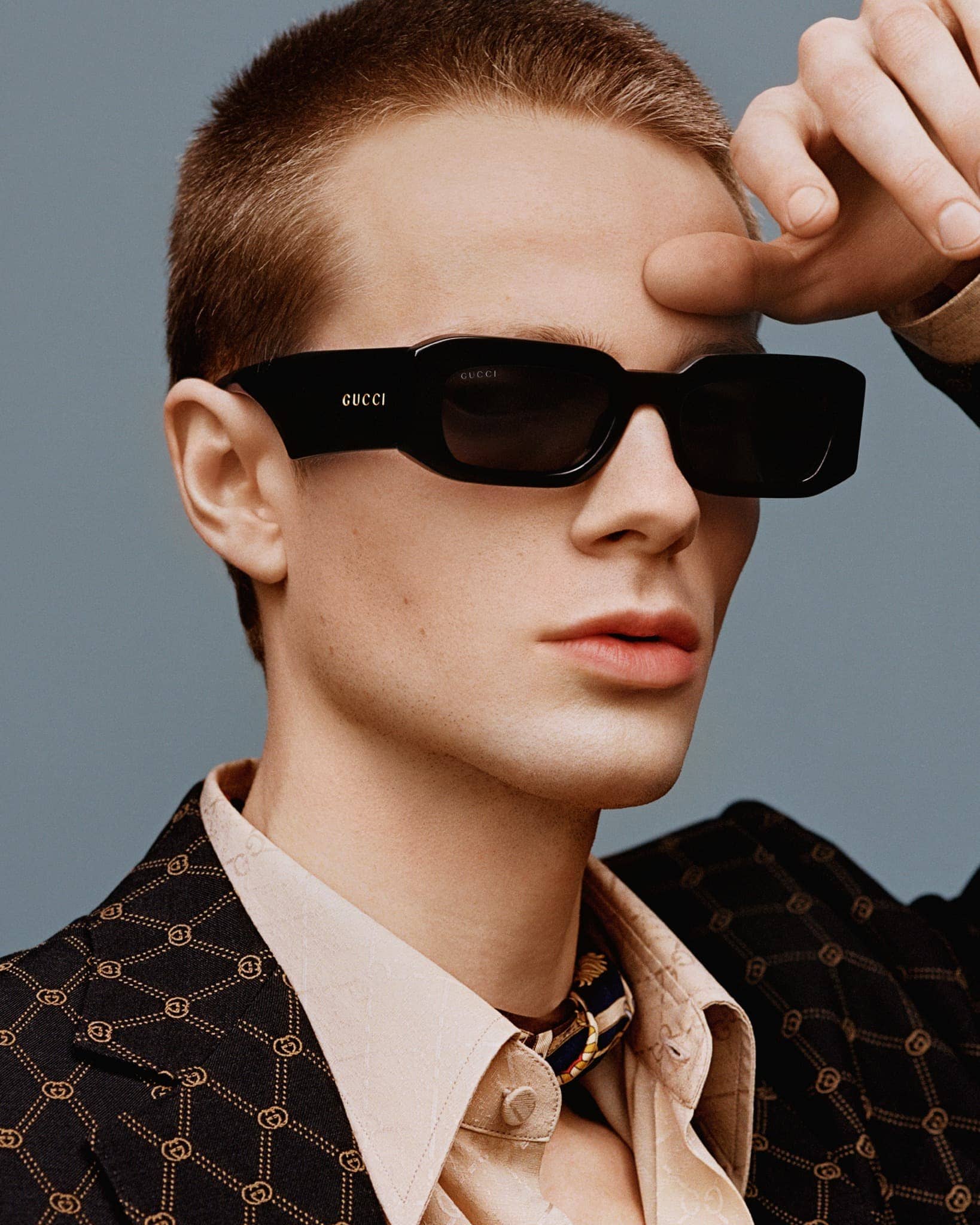 Portrait of a fair-skinned man wearing a patterned blazer and sunglasses with a hand near his forehead in a studio setting.