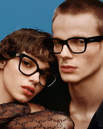 Close-up of a stylish Caucasian couple posing together in eyeglasses, wearing black tops against a solid blue backdrop.