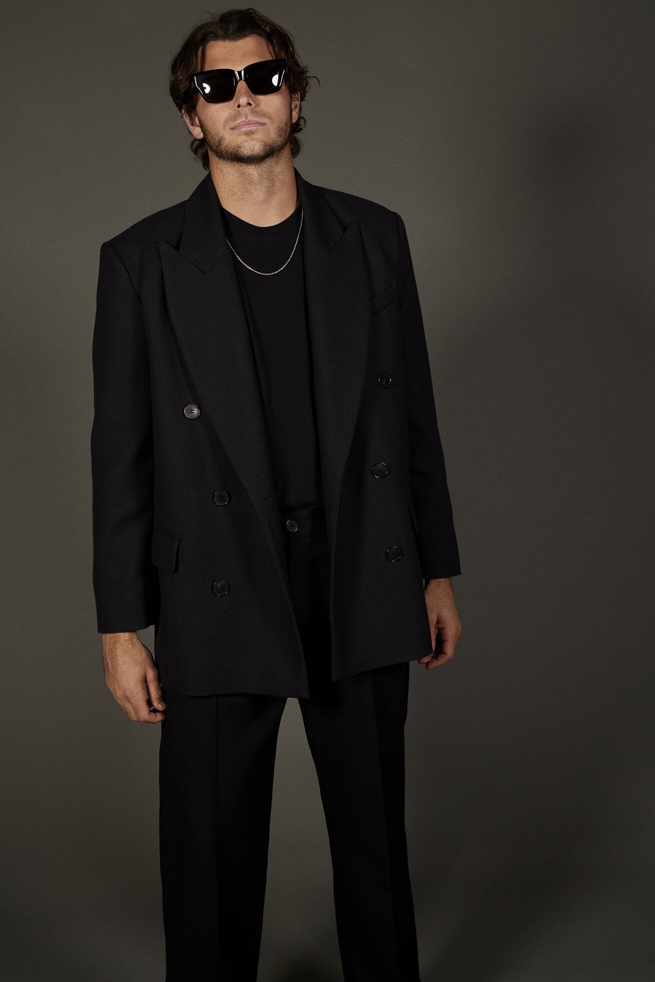 Man wearing a black double-breasted blazer, dark t-shirt, and oversized sunglasses against a neutral dark grey studio background.