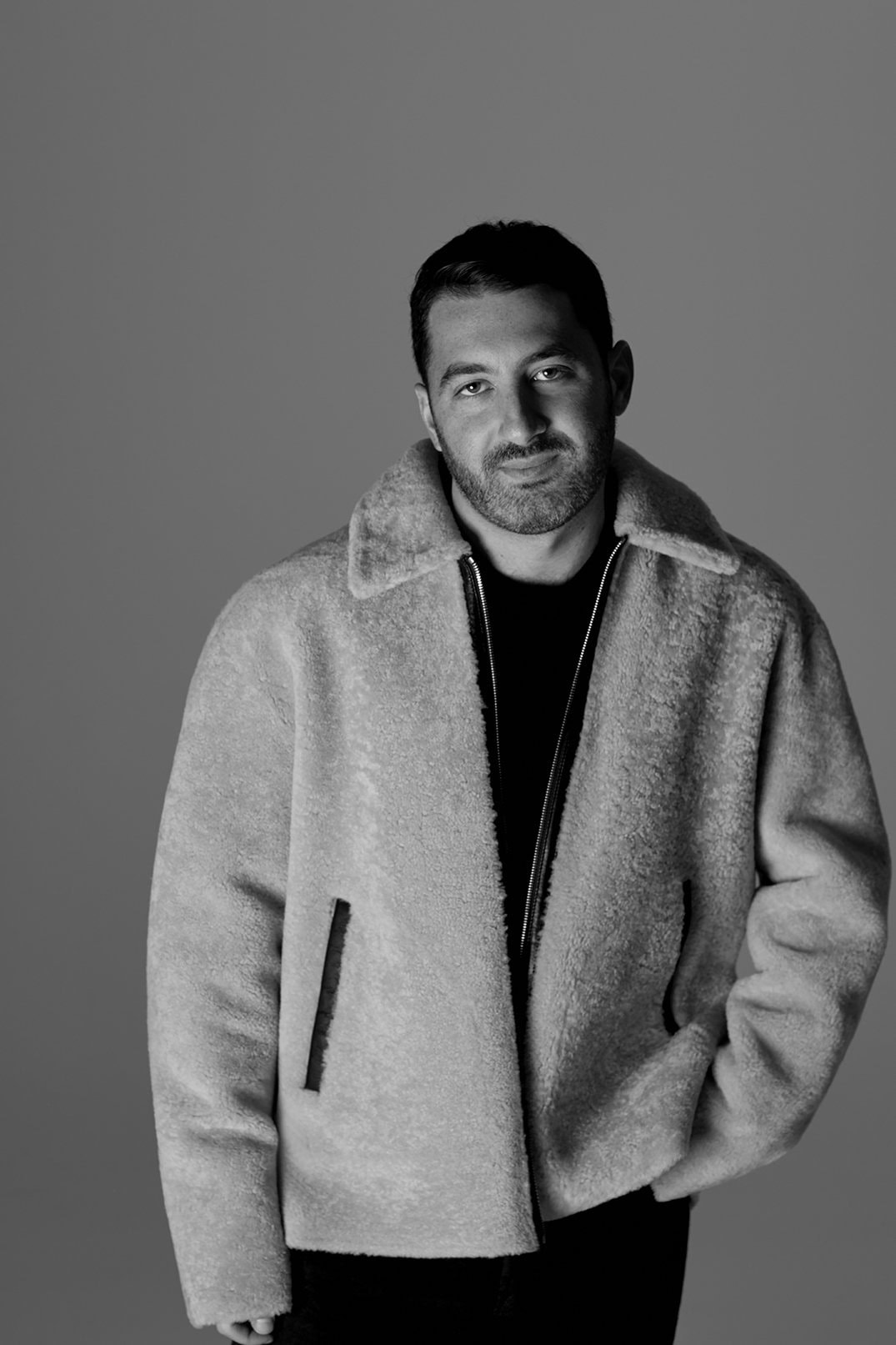 Black and white portrait of an adult male wearing a stylish fuzzy jacket, posing against a neutral background in a studio setting.