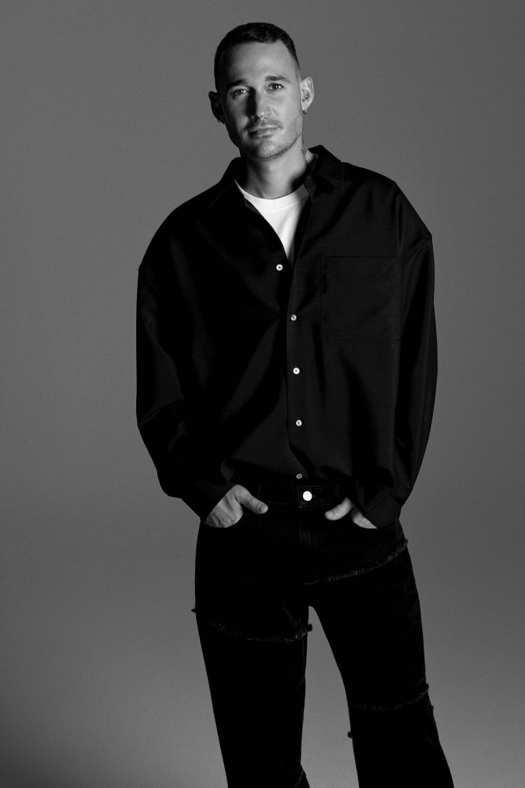 Monochrome studio portrait of a man in a black button-down shirt and dark jeans posing with his hands in his pockets, looking at the camera.