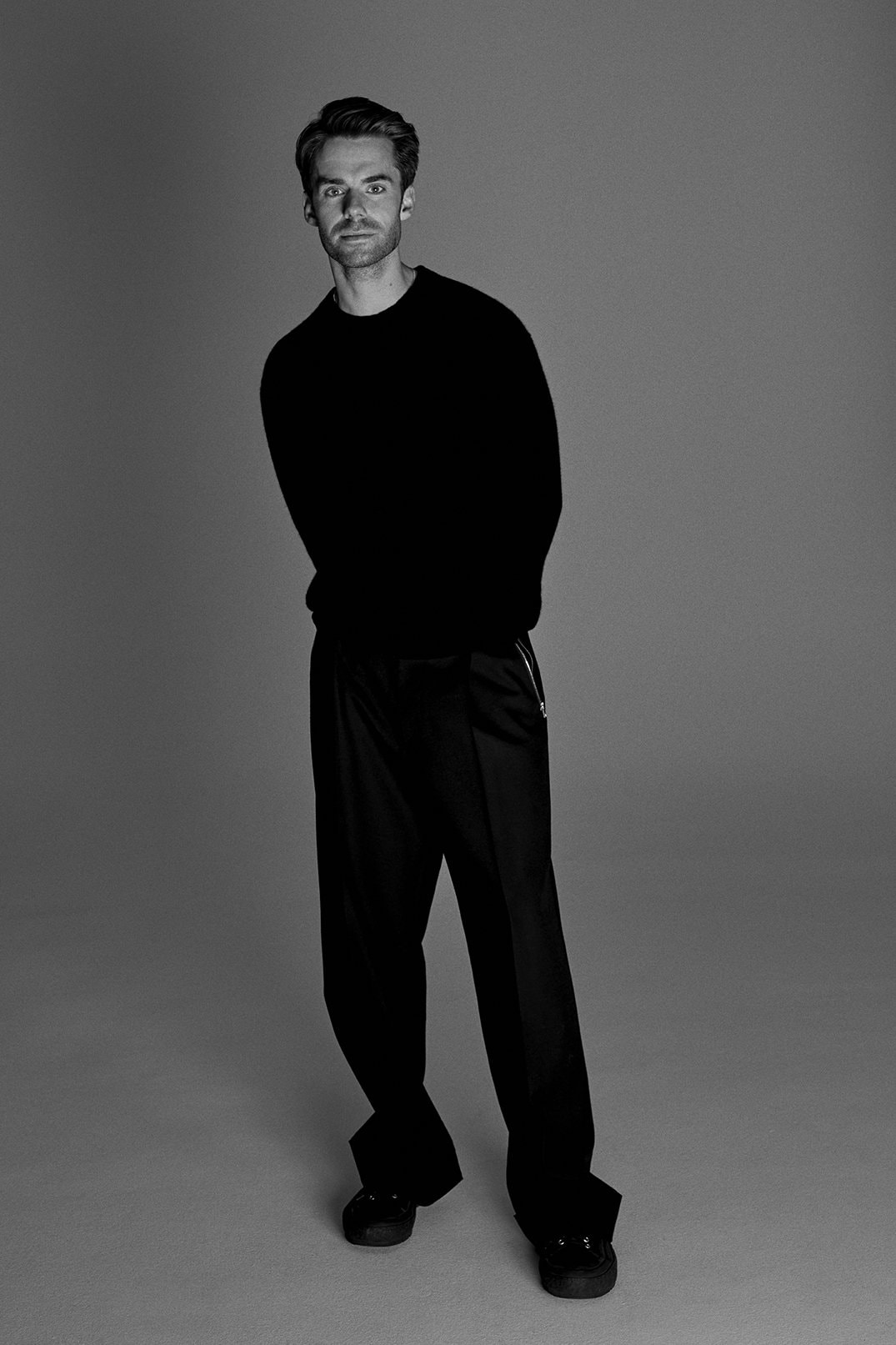Monochrome portrait of a man in a black sweater and trousers standing in a photo studio with neutral grey background and medium shot.