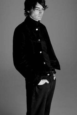 Man posing in a black buttoned jacket and matching dark trousers with hands in pockets against a grey studio backdrop.