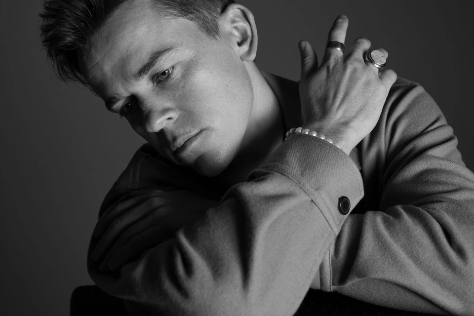 Monochrome portrait of a thoughtful man wearing a suit jacket with jewelry; rings on fingers and pearl bracelet on his wrist.