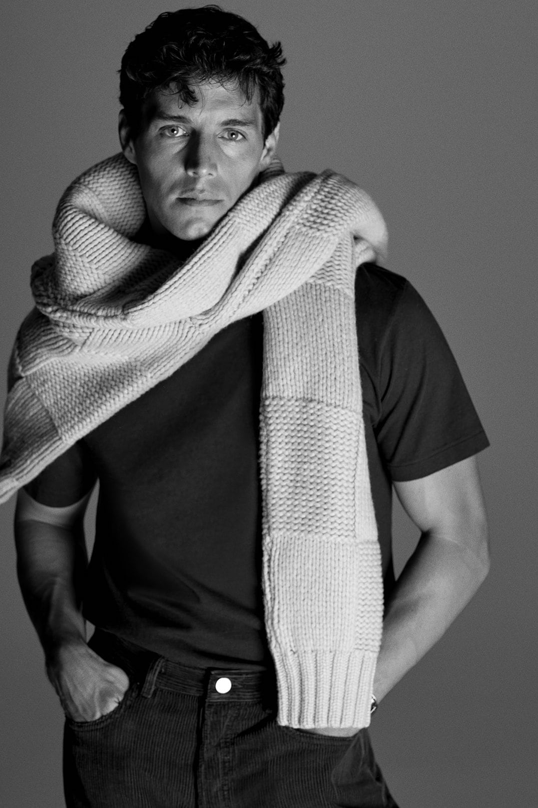 Monochrome studio portrait of a man wearing a dark t-shirt and dark trousers with a chunky textured knit scarf draped around his neck.