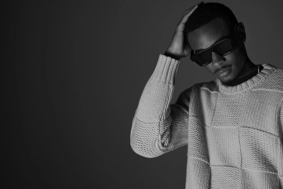 Monochrome portrait of a young adult male with sunglasses and a sweater, his hand on his head, against a dark gray backdrop.