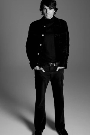Monochrome portrait of a man wearing a dark jacket and trousers standing against a grey backdrop for FRAME