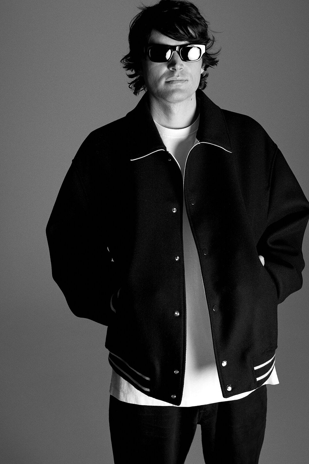 A man with dark hair and sunglasses stands in front of a grey background, wearing a black letterman jacket and a white t-shirt.