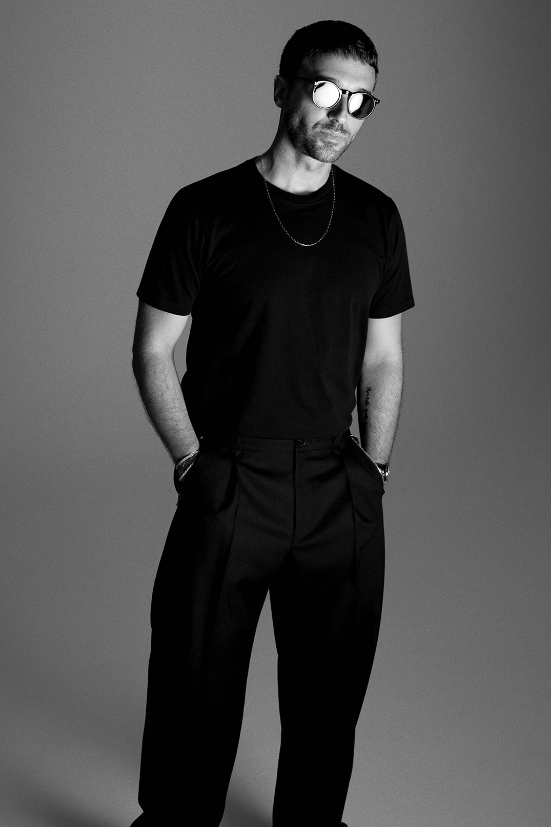 Black and white studio shot of a man wearing a black t-shirt, black pants, and sunglasses, standing against a gray backdrop.
