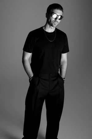 Black and white studio shot of a man wearing a black t-shirt, black pants, and sunglasses, standing against a gray backdrop.