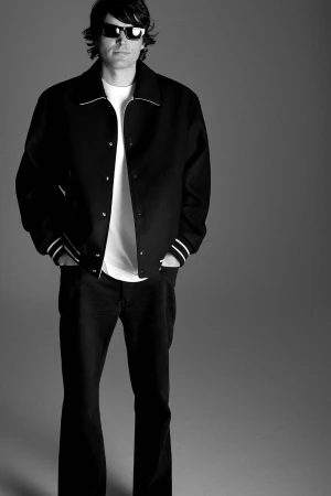 Full-length studio shot in black and white of an adult male wearing sunglasses, black letterman jacket, white t-shirt, and dark denim jeans.