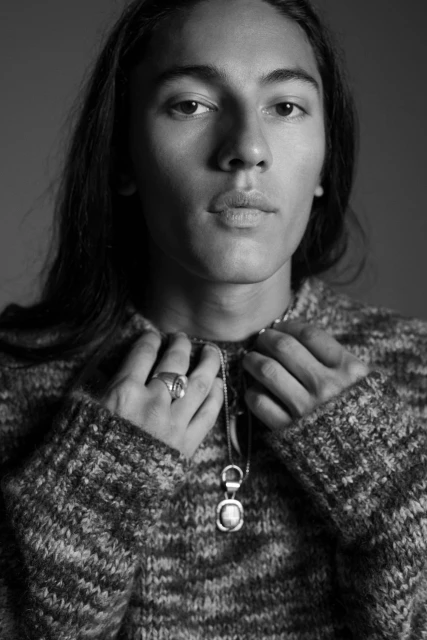 Black and white portrait of a young adult male wearing a knit sweater and a pendant necklace, gazing directly at the camera.