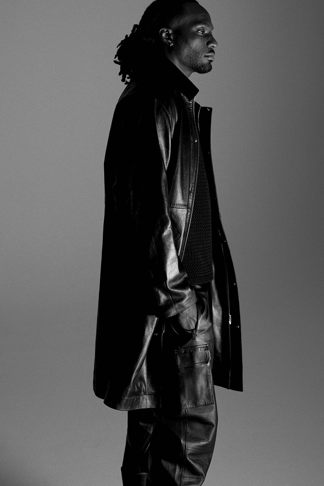 Monochrome portrait of a Black adult male in a leather coat and pants, posing in profile against a plain gray background in studio light.
