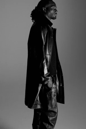 Monochrome portrait of a Black adult male in a leather coat and pants, posing in profile against a plain gray background in studio light.