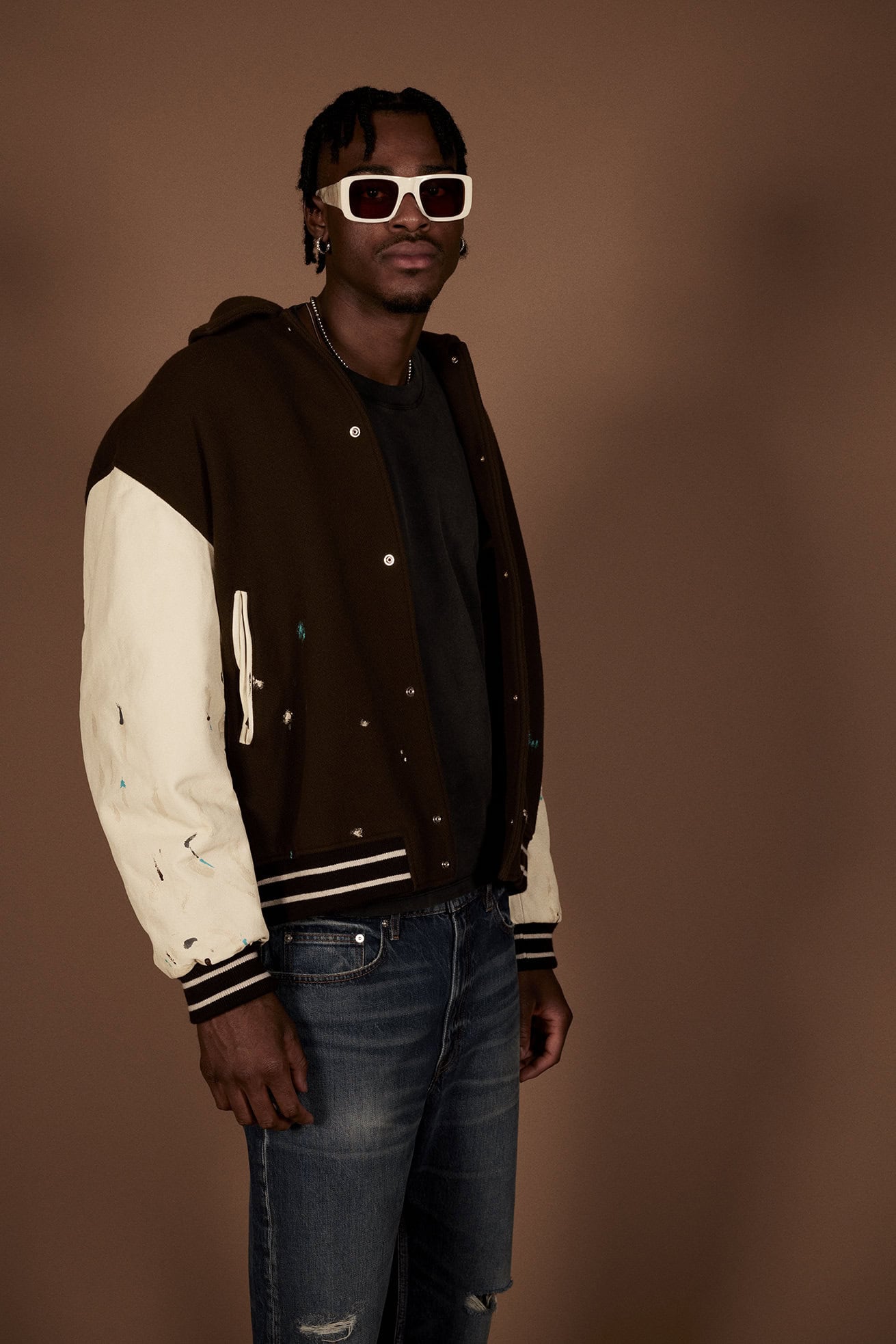 A man with braided hair wearing white sunglasses, a dark brown and off-white letterman jacket, and blue jeans against a brown background.