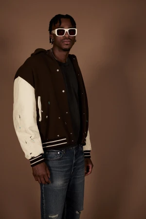 A man with braided hair wearing white sunglasses, a dark brown and off-white letterman jacket, and blue jeans against a brown background.