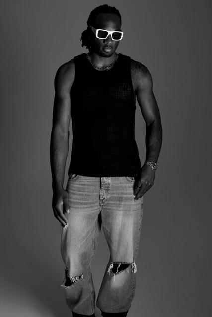 Black and white studio shot of a Black man wearing a tank top, distressed denim, white-frame glasses and wrist watch, looking forward.