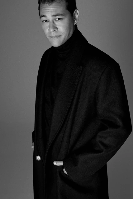 Black and white portrait of a man wearing a dark coat and turtleneck, posing indoors with his hands in his pockets. Fashion.